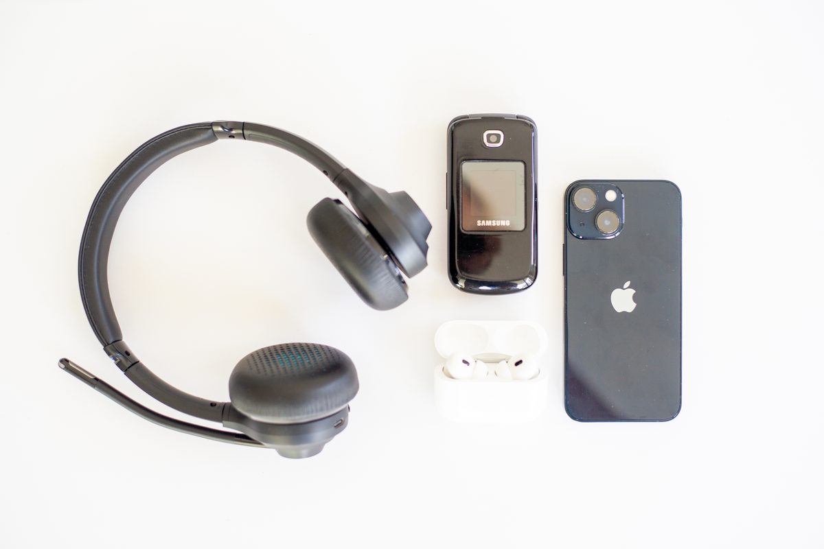 A headset, a flip phone, earbuds and an iPhone laid out on a white desk