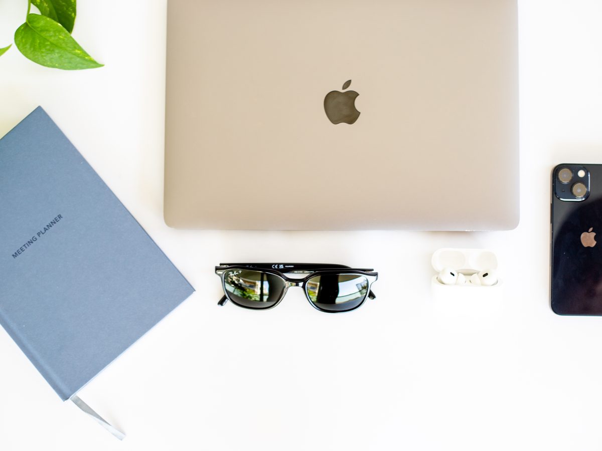 A notebook, laptop, sunglasses, earbuds and a phone on a white desk